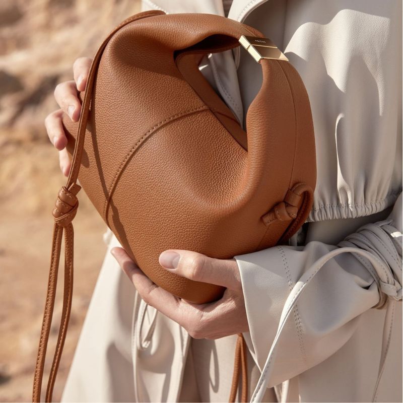 Brown leather handbag held by a person wearing a light-colored outfit.