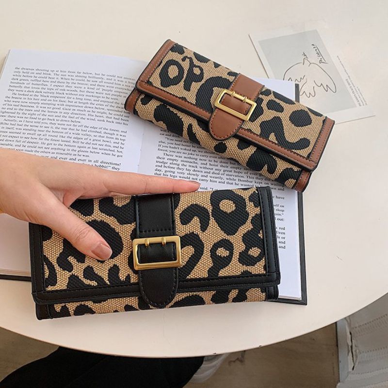Leopard print wallet with gold buckle held by a hand on a table with an open book.
