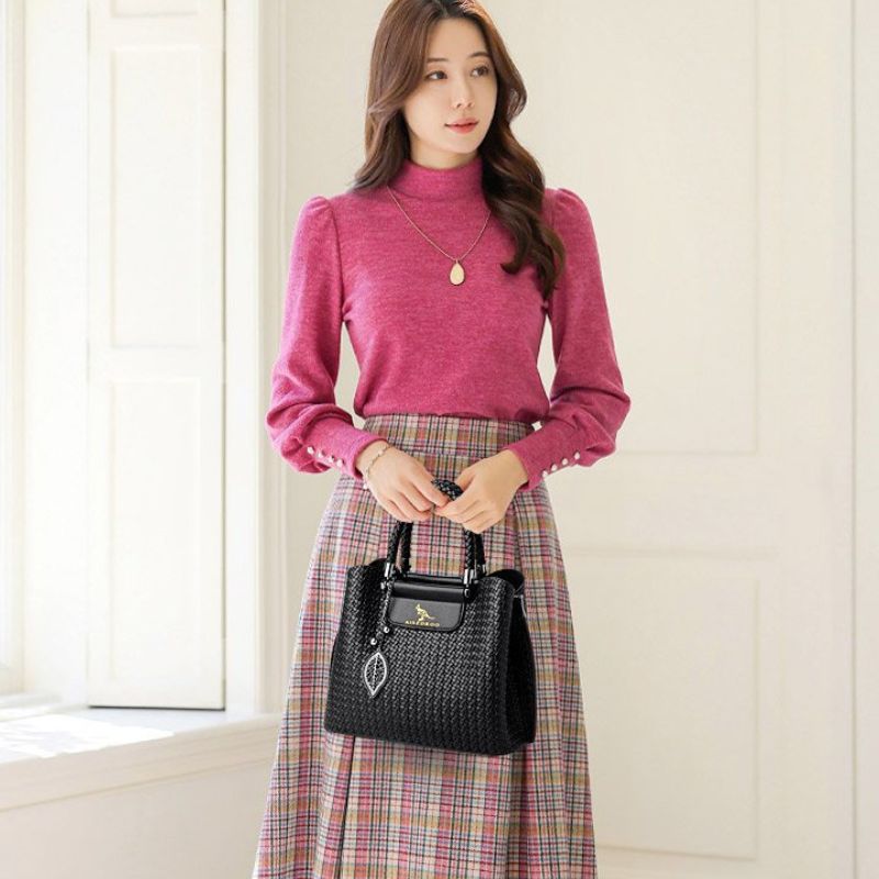 Woman wearing a pink sweater and plaid skirt holding a black handbag indoors.