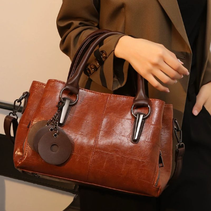 Brown leather handbag with decorative tag held by a person wearing a brown coat.