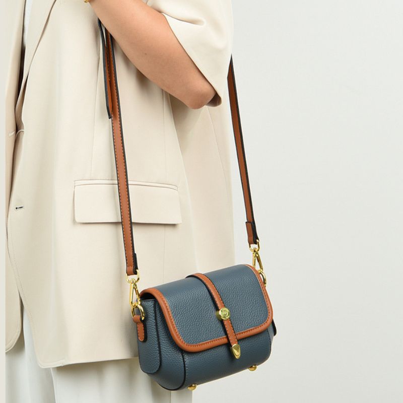 Blue handbag with brown accents held by a person wearing a beige outfit on a light gray background