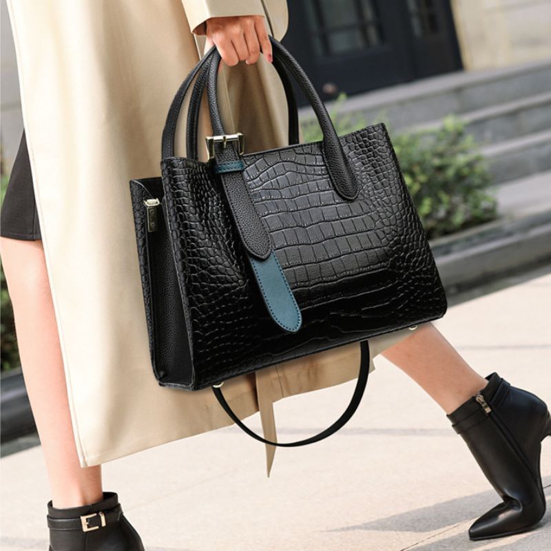 Person holding a black crocodile-patterned handbag with a blue strap outdoors.